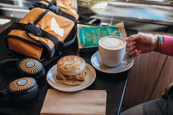 Close up of hand on coffee beverage with breakfast sandwich, headphones, work bag, and book on cafe table