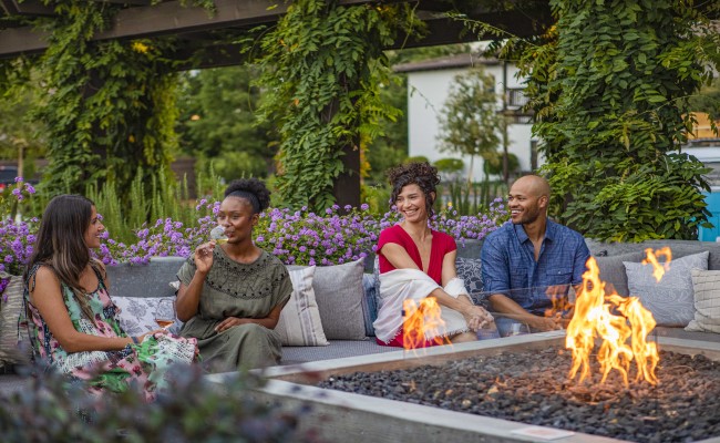 Wit and Wisdom exterior. Friends enjoying glass of wine outdoors in front of fire pit with arbors in the background cover in vines and flowers.