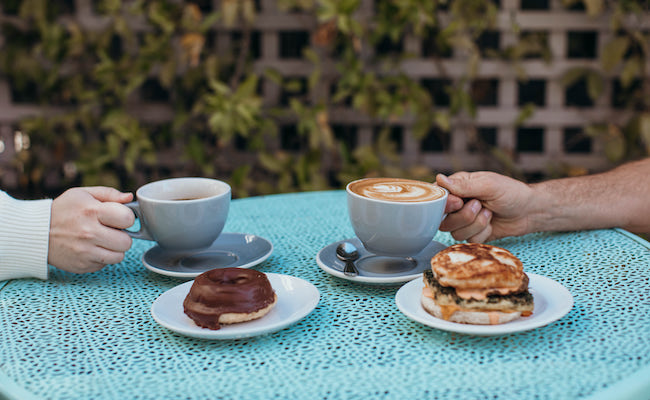 Retrograde Coffee Roasters. Vegan doughnut, breakfast sandwich, and two coffee lattes on an outdoor cafe table with foliage in the background. Hand holding handle of each cup.